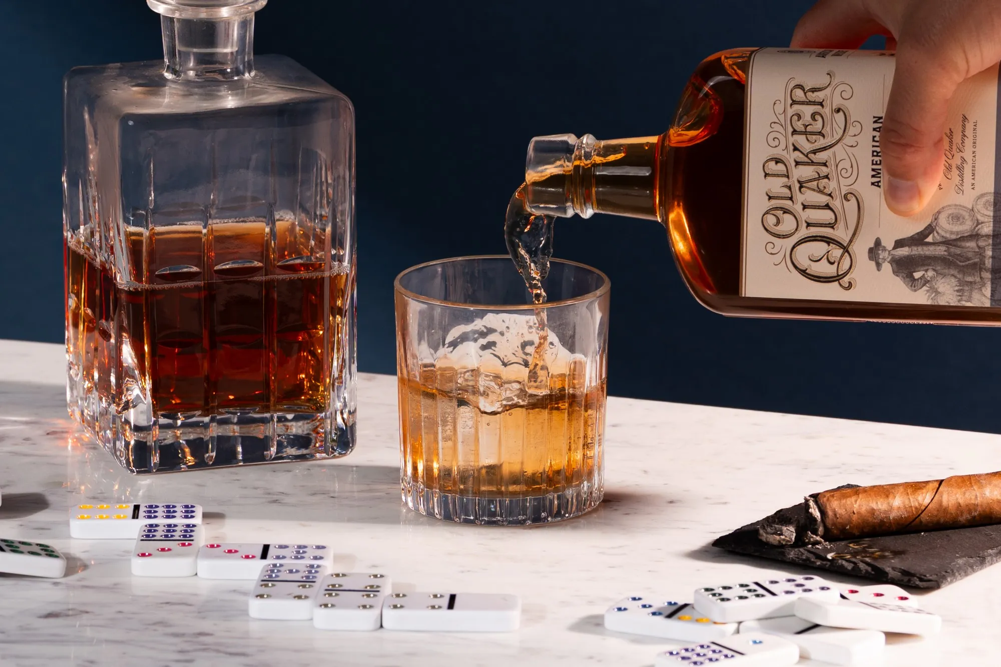 Old Quaker Straight American Whiskey poured into a glass beside a crystal decanter, dominoes and a cigar